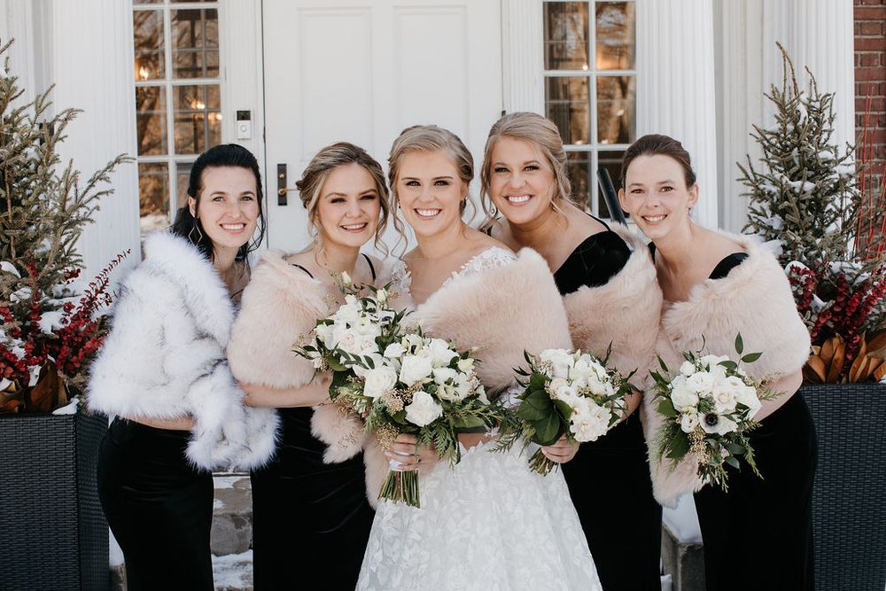 Smiling bride and four bridesmaids in black dresses with fur wraps holding white bouquets outdoors in snow.