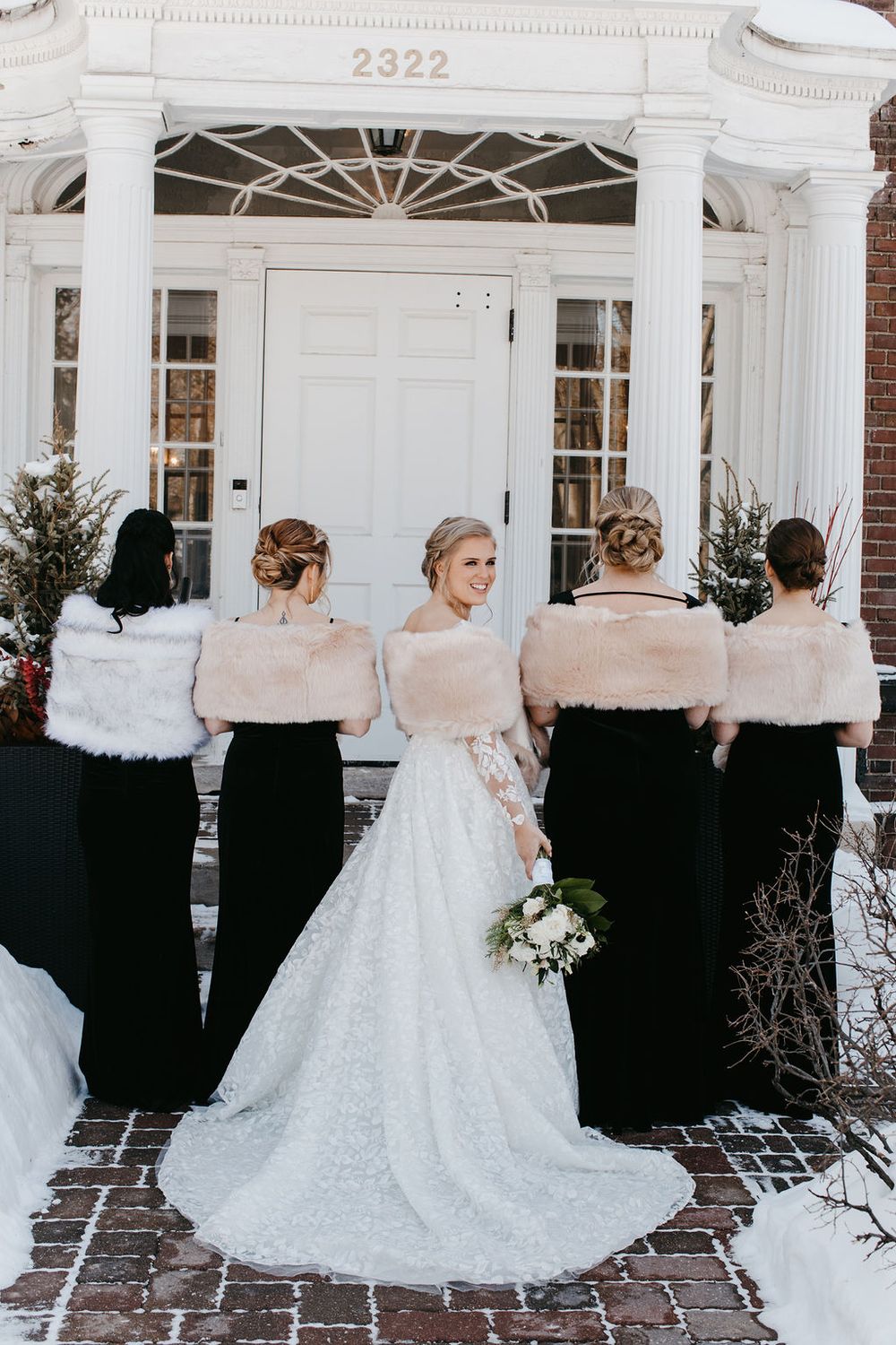 Bride in white lace gown facing door with four bridesmaids in black dresses and fur wraps at building 2322.