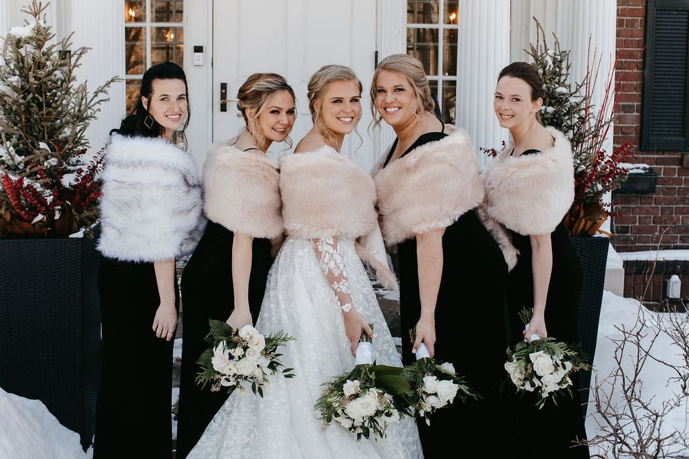 Bride in white lace dress smiling with four bridesmaids wearing black gowns and fur wraps holding bouquets.