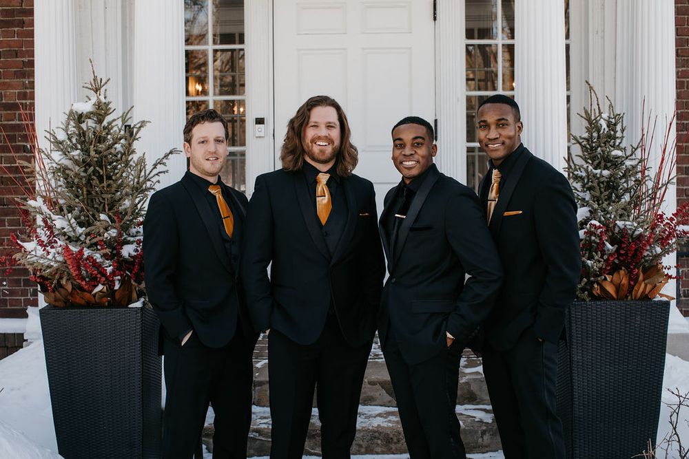 Four groomsmen in black tuxedos with gold ties posing together outside a brick building in winter.