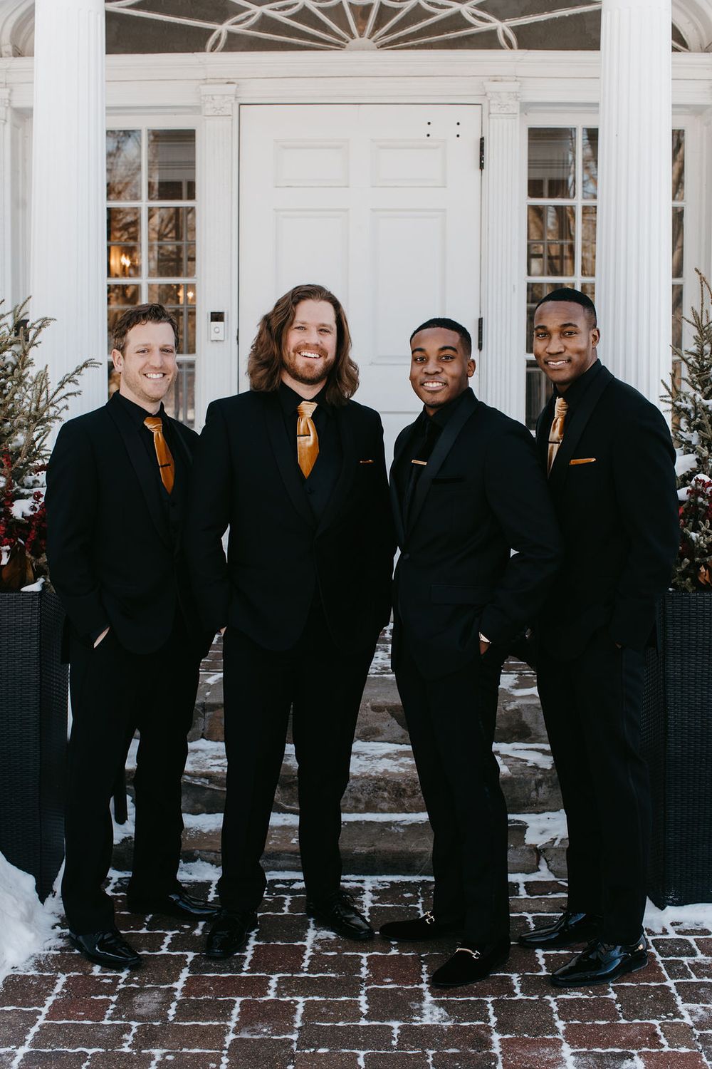 Four groomsmen in black suits and gold ties posing formally outside a white-door brick building in winter.