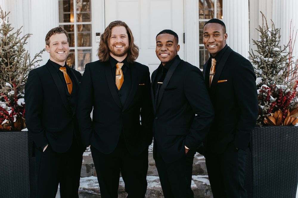 Four groomsmen pose together in matching black suits with gold ties outside a white wedding venue.