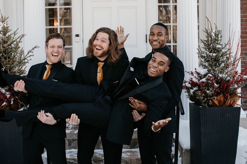 Four groomsmen in black tuxedos with gold ties laugh as one is lifted up outside a white building.