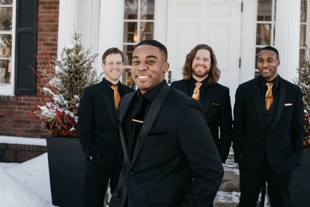 Smiling groom in black tuxedo stands in front of three groomsmen outside a brick building in winter.