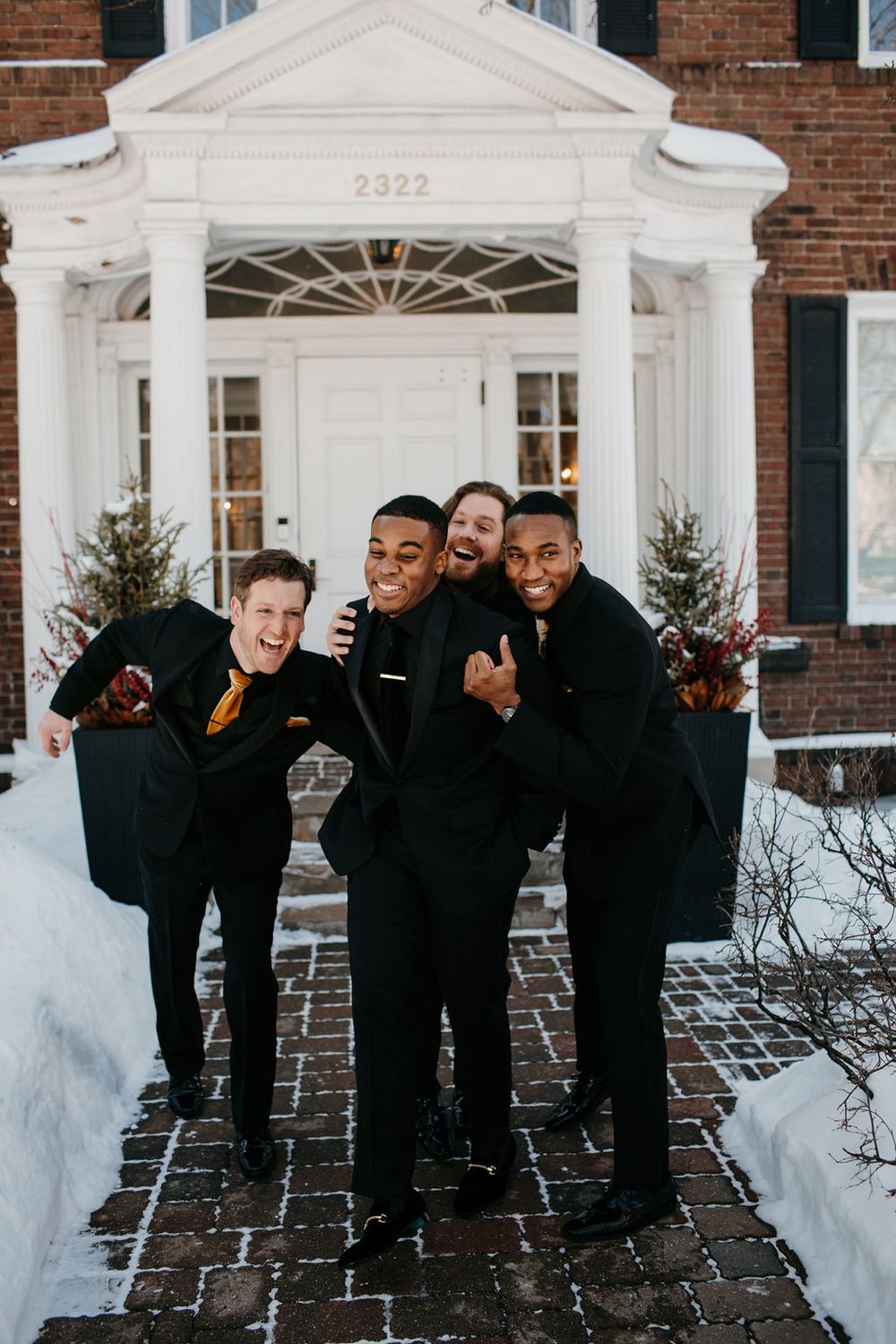 Four groomsmen in black suits laughing outside a brick building in winter snow.