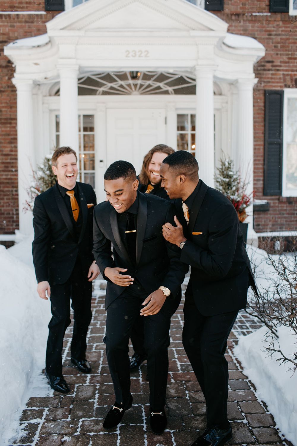 Four groomsmen in black tuxedos with gold ties laughing outside a brick building in winter snow.