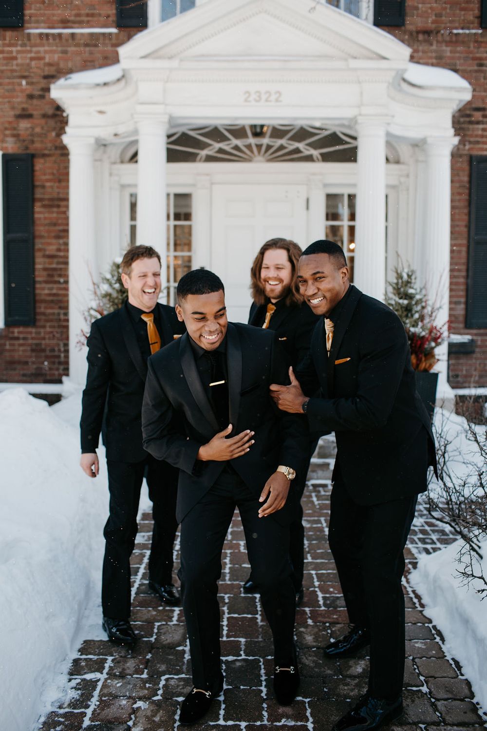 Four groomsmen in black suits with gold ties smiling and posing outside a brick building numbered 2322.
