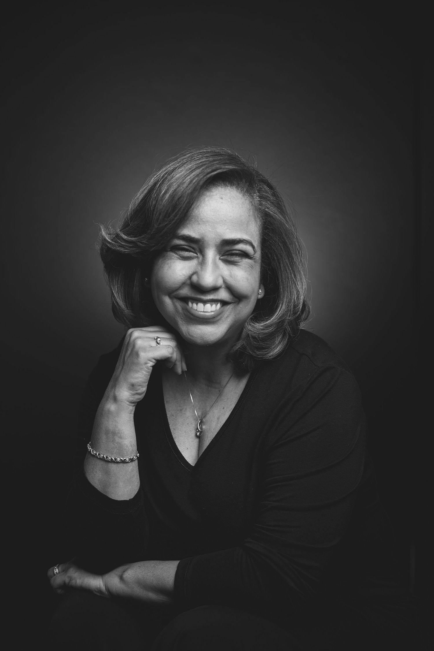 Black and white portrait of a smiling middle-aged woman in black top, resting chin on hand.