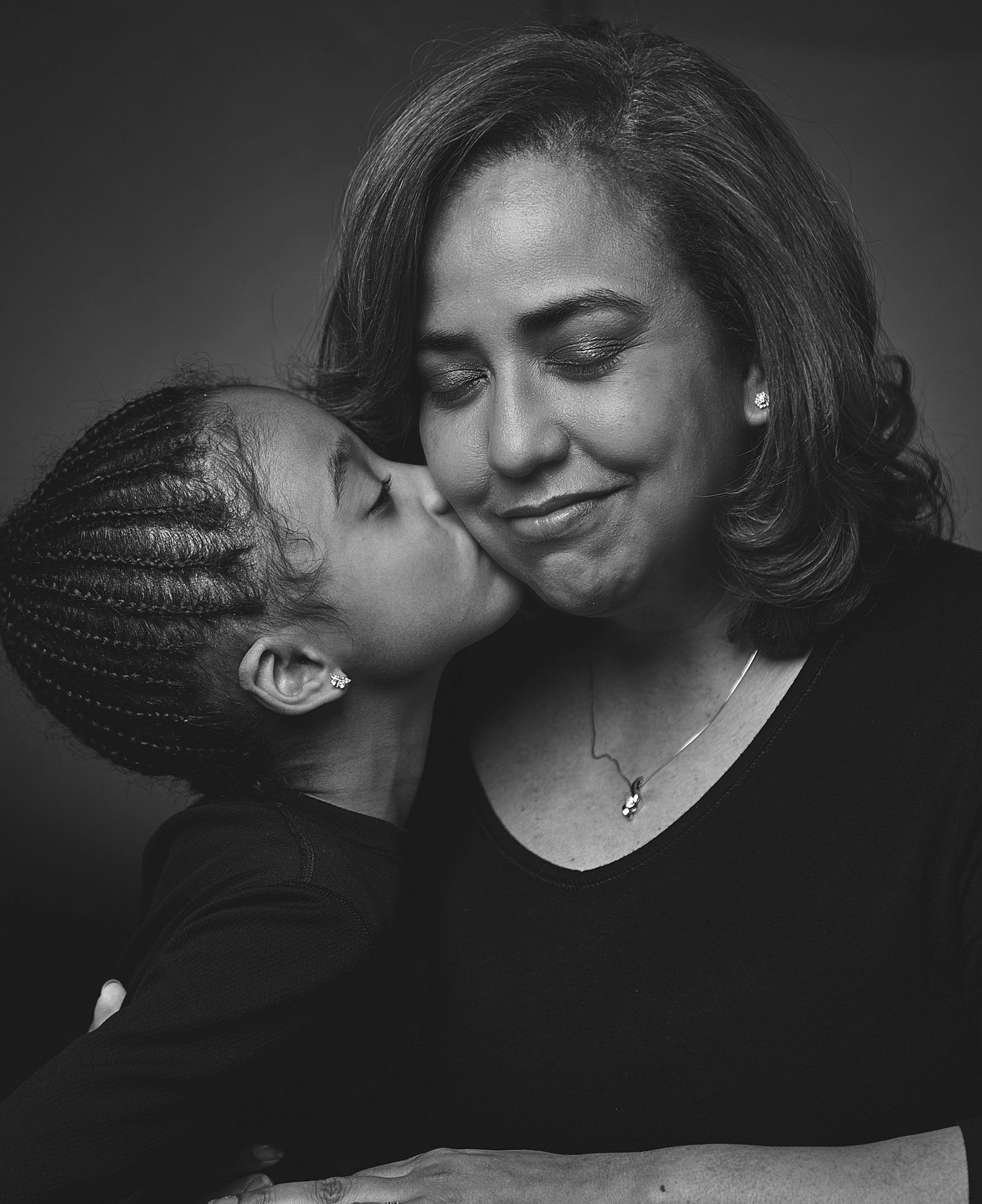 Black and white image of a young girl with braids giving a woman a kiss on the cheek.