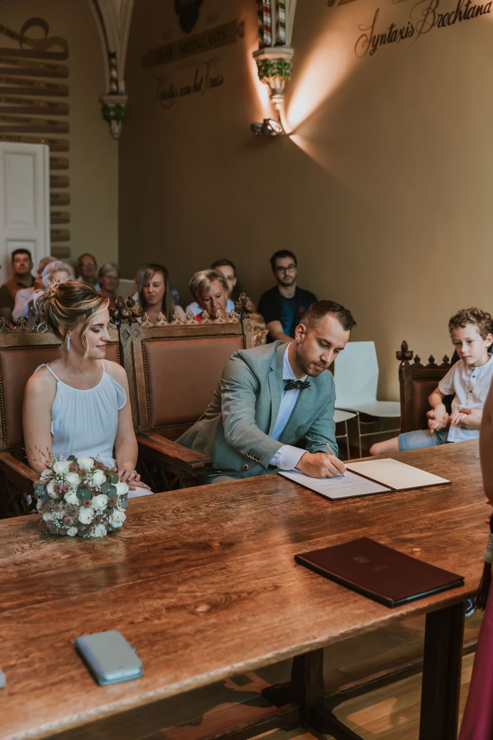 A couple sits at a wooden table with bouquet during document signing in an ornate room.