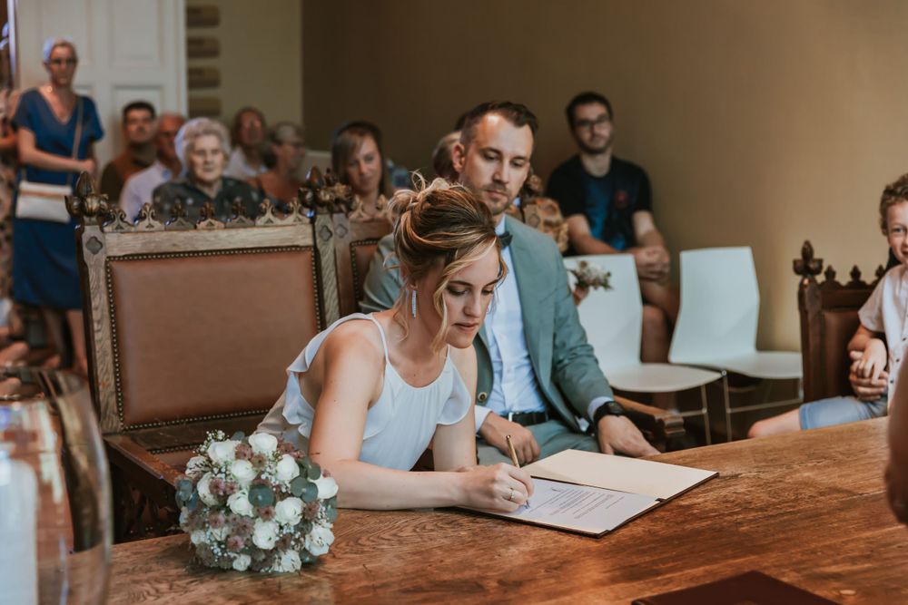 A couple signs documents at a wooden table in a ceremonial room with guests seated behind.