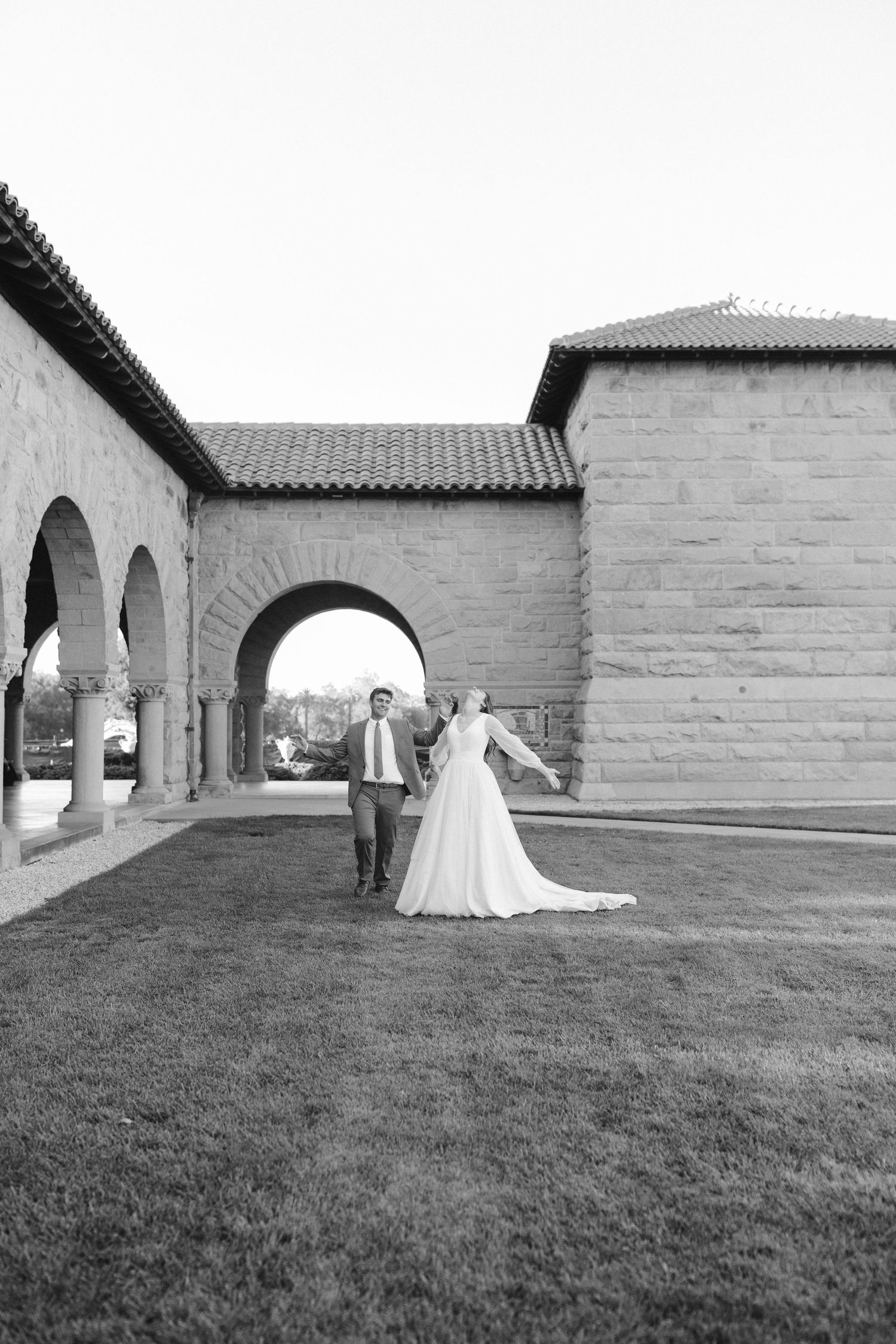 A wedding couple poses on grass near Mediterranean architecture with arched walkways.