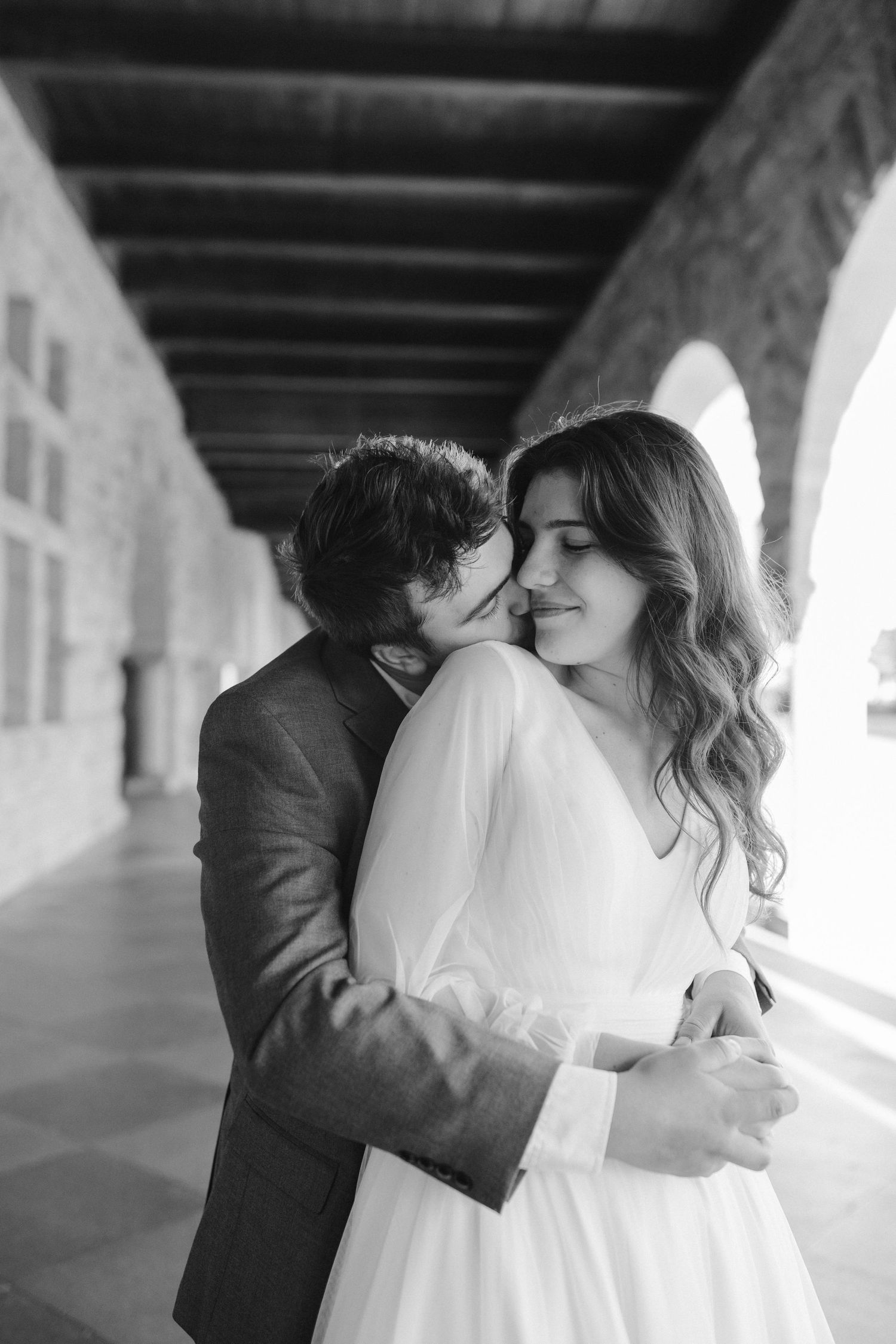 Black and white intimate moment between a couple in wedding attire under historic arches.