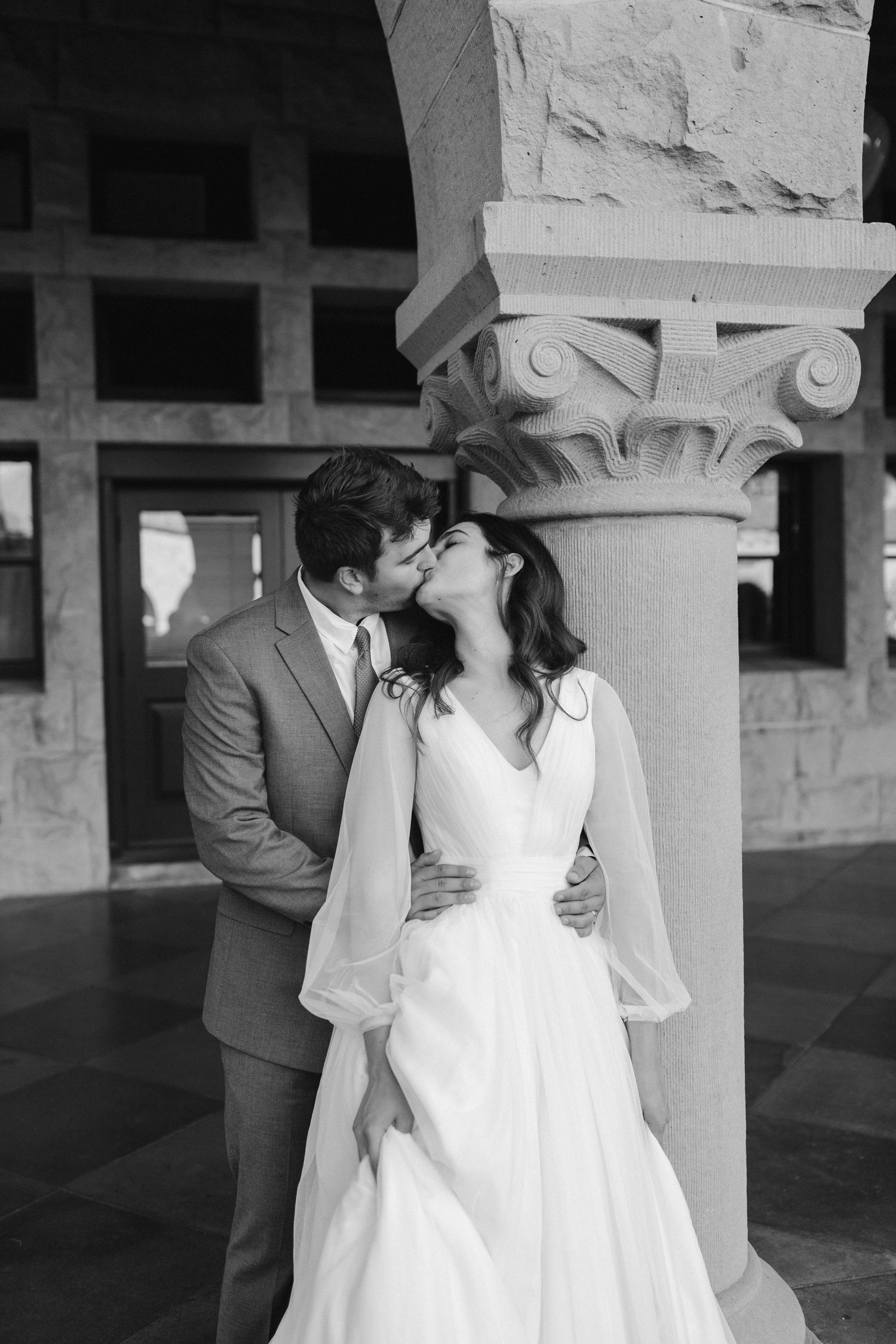 A romantic wedding couple shares a kiss beside an ornate stone column in a black and white photo.