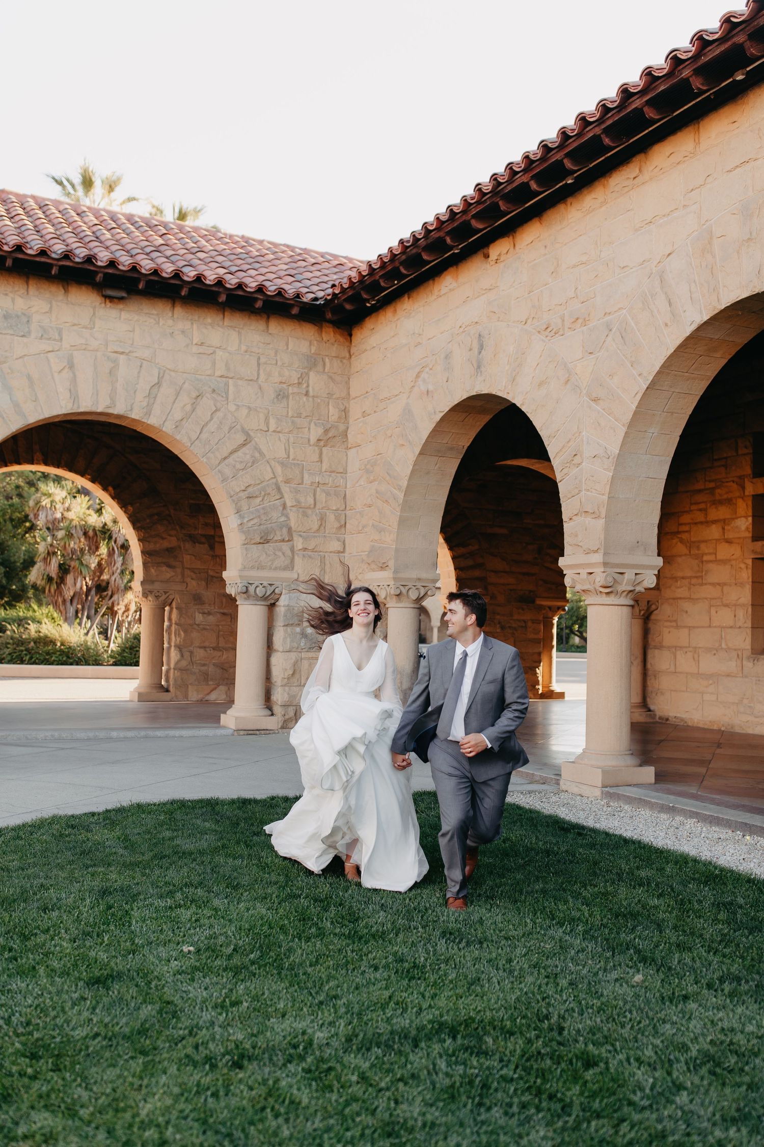 A joyful wedding couple walks under Mediterranean arches with red tile roof and grass.