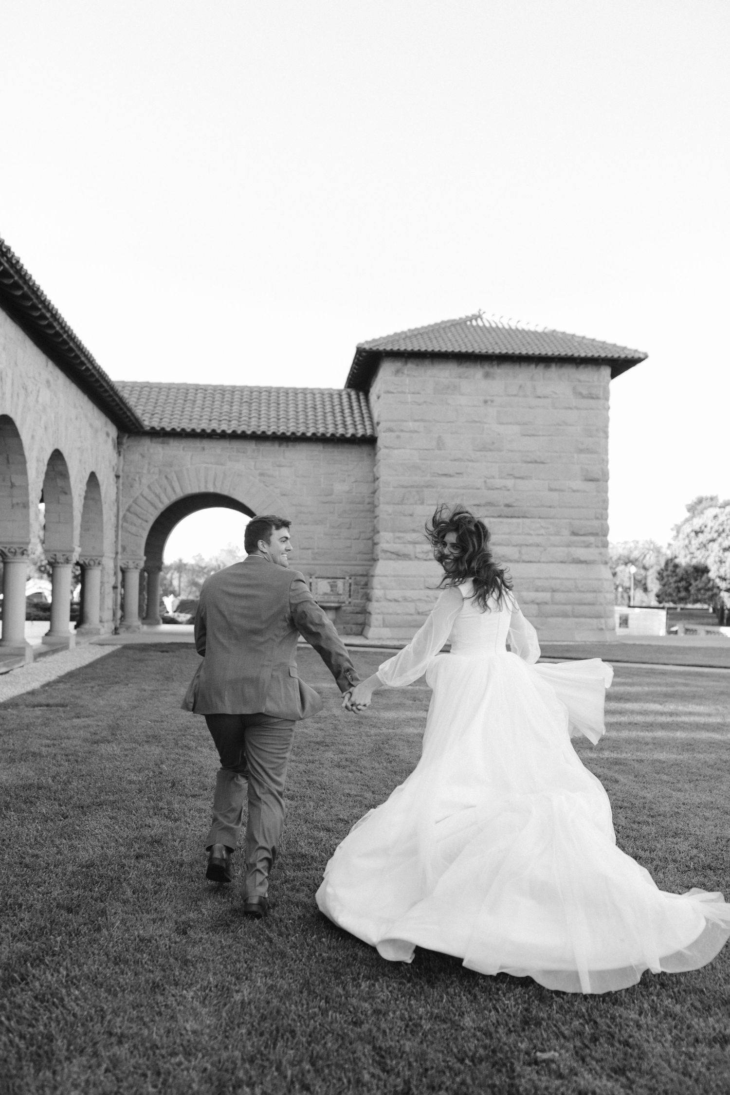 A black and white photo of newlyweds running along an arched colonnade pathway.