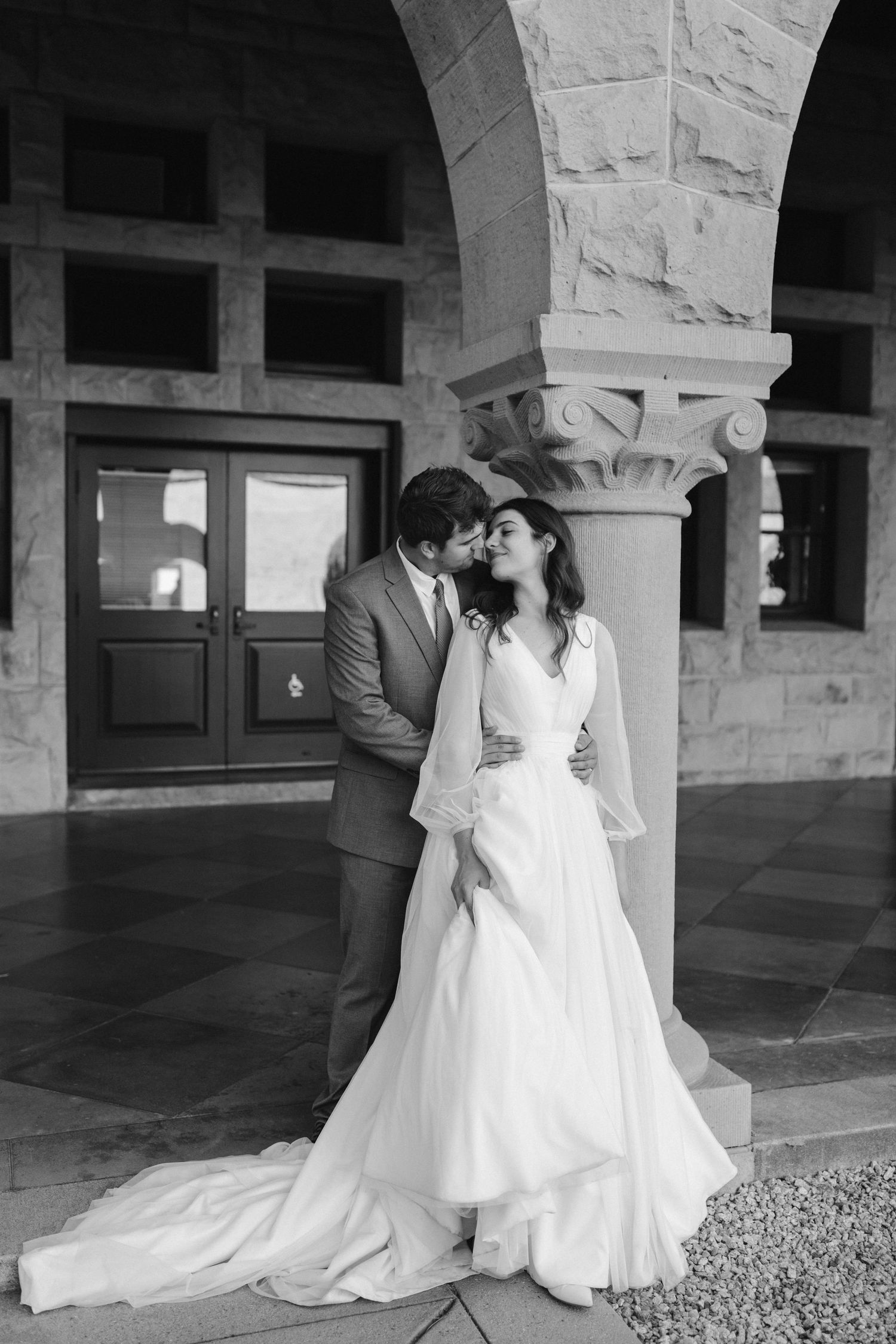 Black and white portrait of a couple sharing an intimate moment by an ornate stone column.
