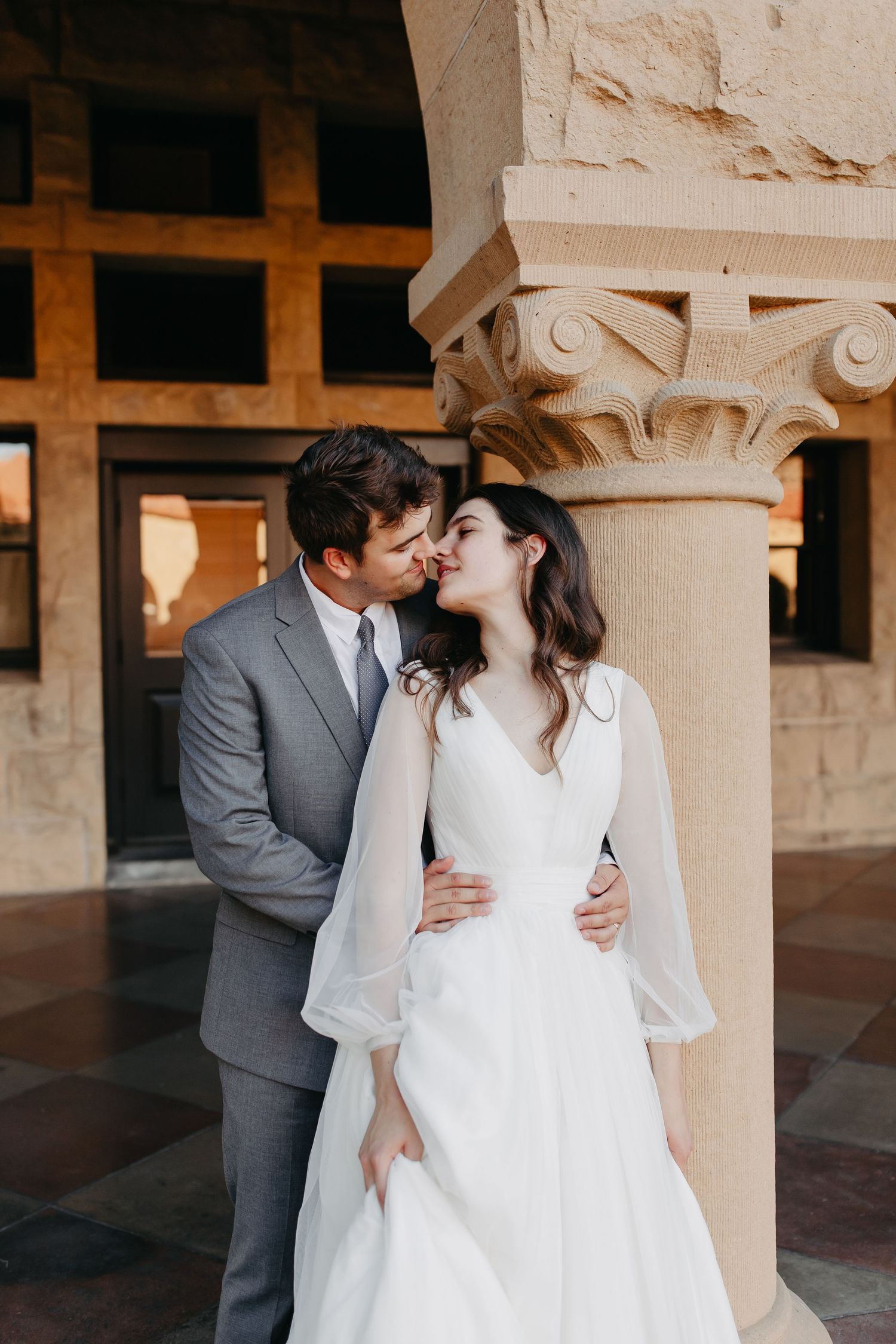 A romantic wedding portrait of a couple embracing by a decorative stone column in warm sunlight.