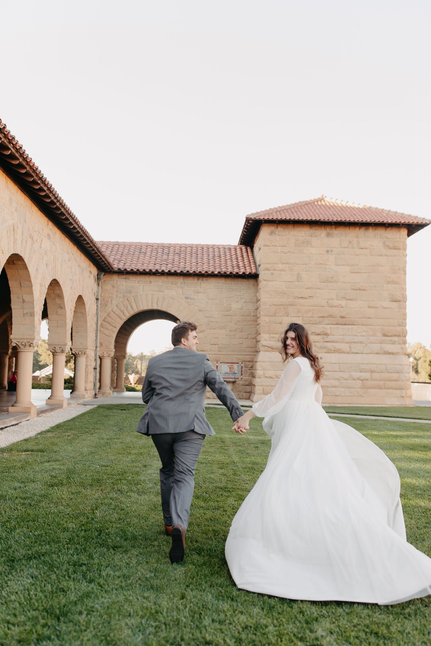 A wedding couple runs playfully across grass near a Spanish-style building with arched walkways.