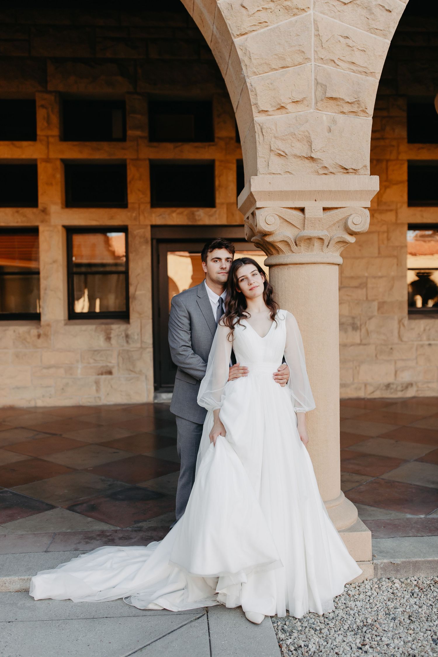 A wedding couple poses elegantly against a stone pillar in Mediterranean-style architecture.