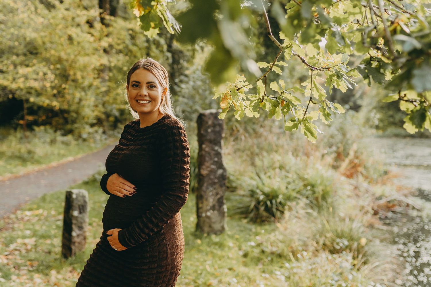 Expectant mother in black dress standing in sunlit garden path.