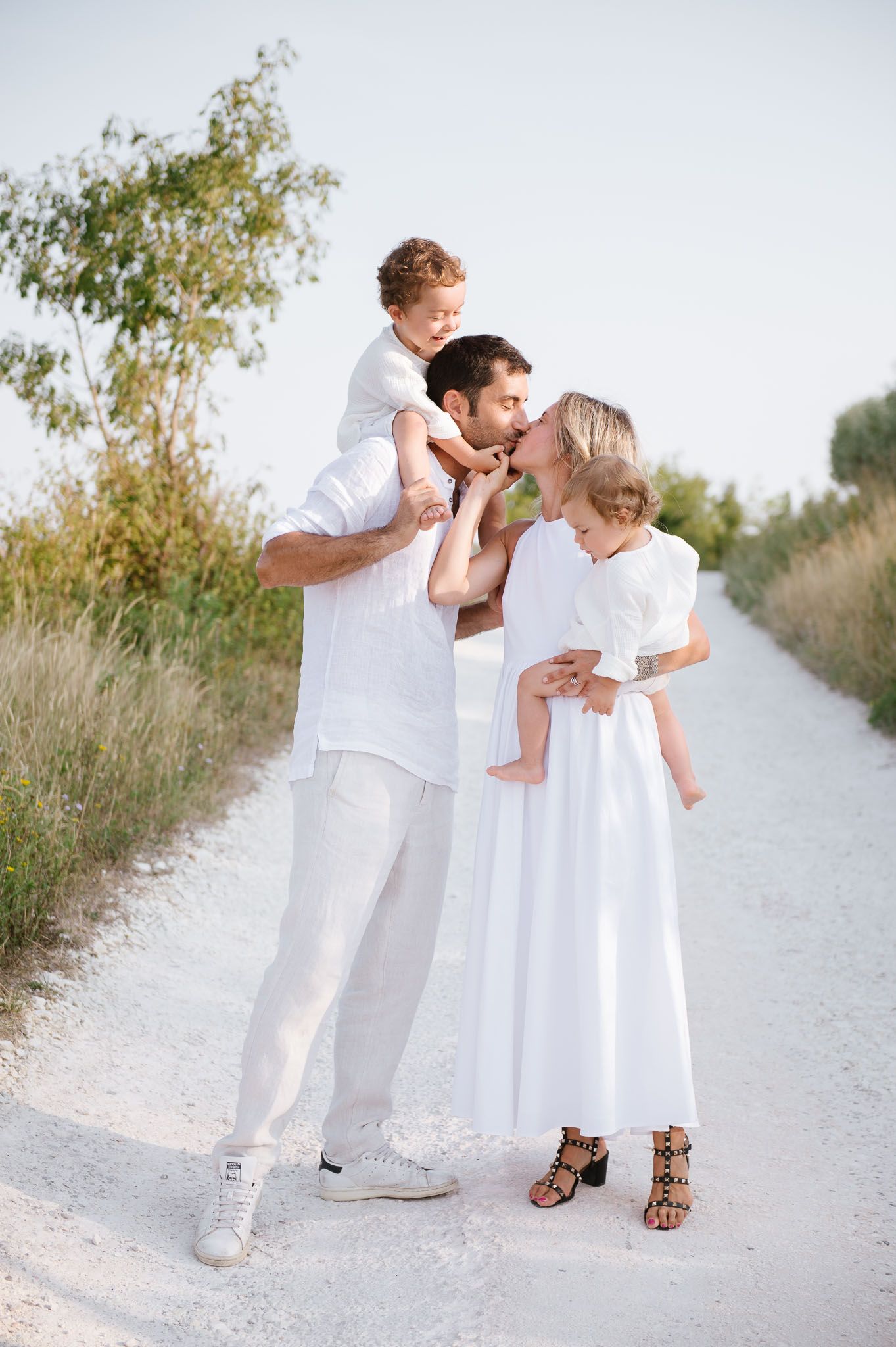 A family in white clothing poses together on a sunlit path surrounded by nature.