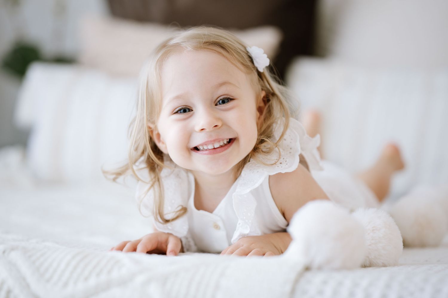 A child in white lies on a white bed with a small bunny toy.
