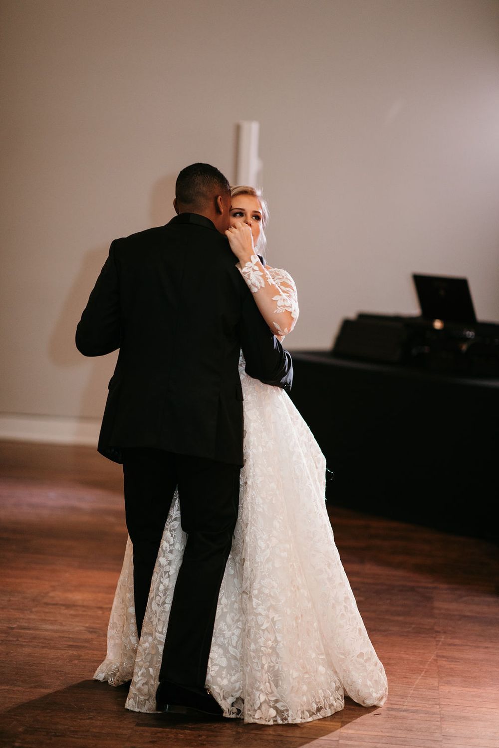 Bride wiping tears while sharing an emotional first dance with her groom in a lace wedding dress.