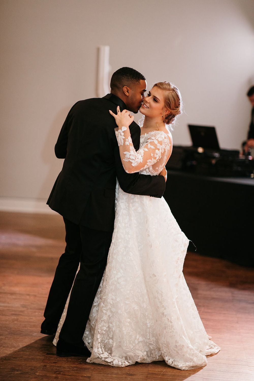 Interracial bride and groom sharing their first dance, with the bride smiling in a lace long-sleeve gown.