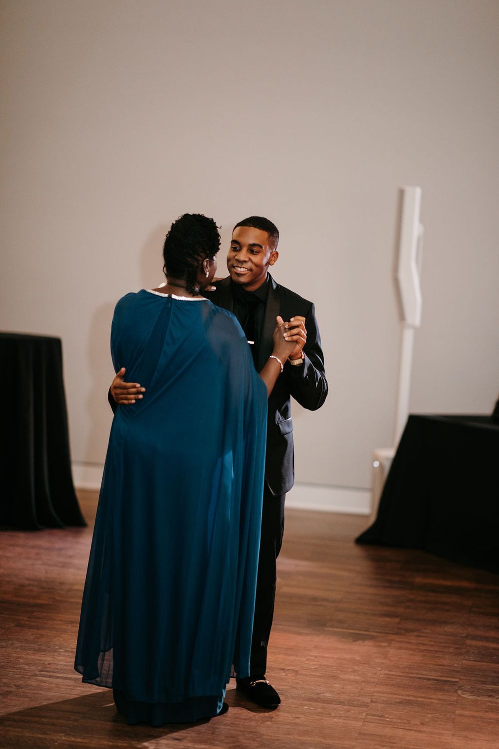 Groom smiling while dancing with his mother in teal dress at wedding reception.