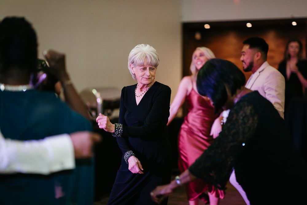 Elderly woman in black outfit energetically dancing at a wedding reception with guests around her.