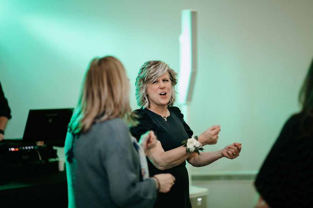 Woman in black outfit dancing enthusiastically under green lights at wedding reception.