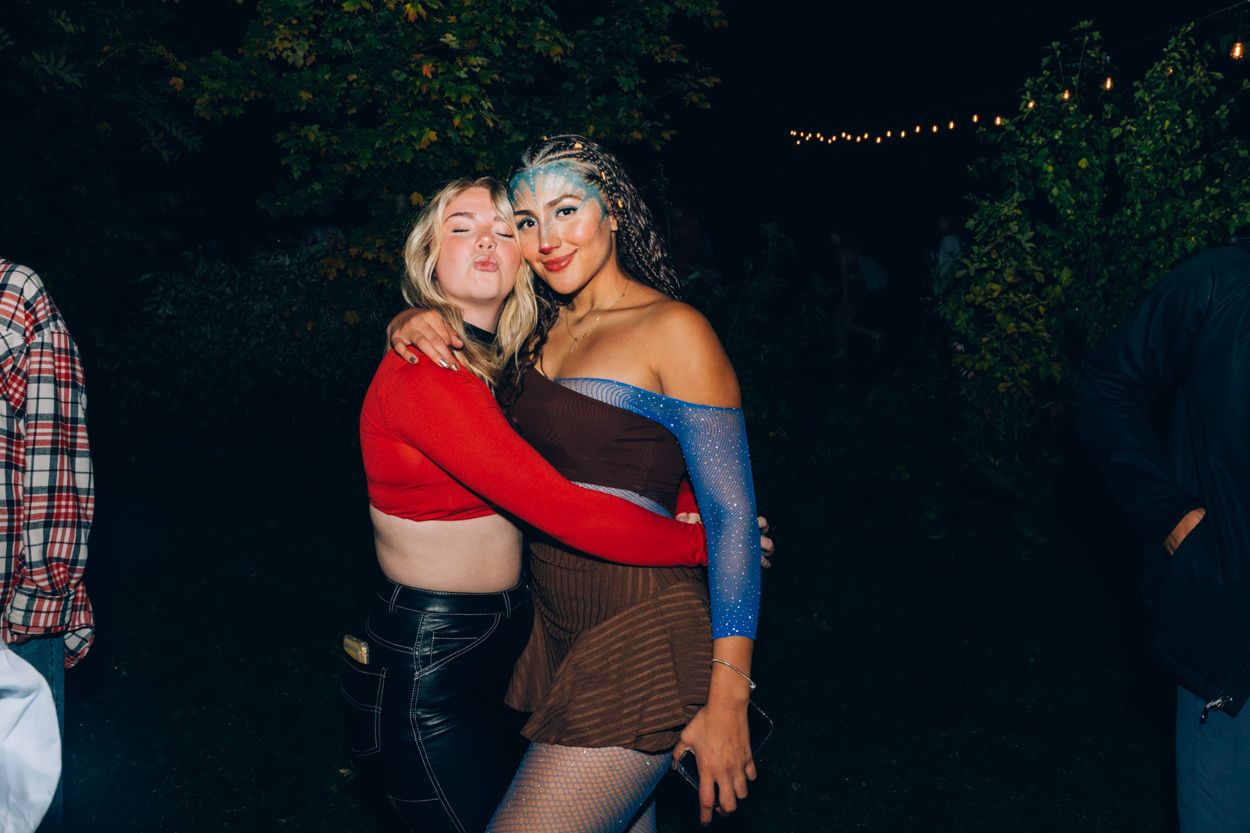 Two people in party outfits embrace while posing at nighttime outdoor gathering.
