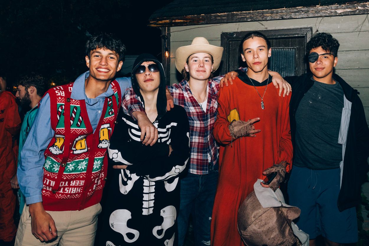 Group wearing various costumes including skeleton outfit and cowboy hat at Halloween party.