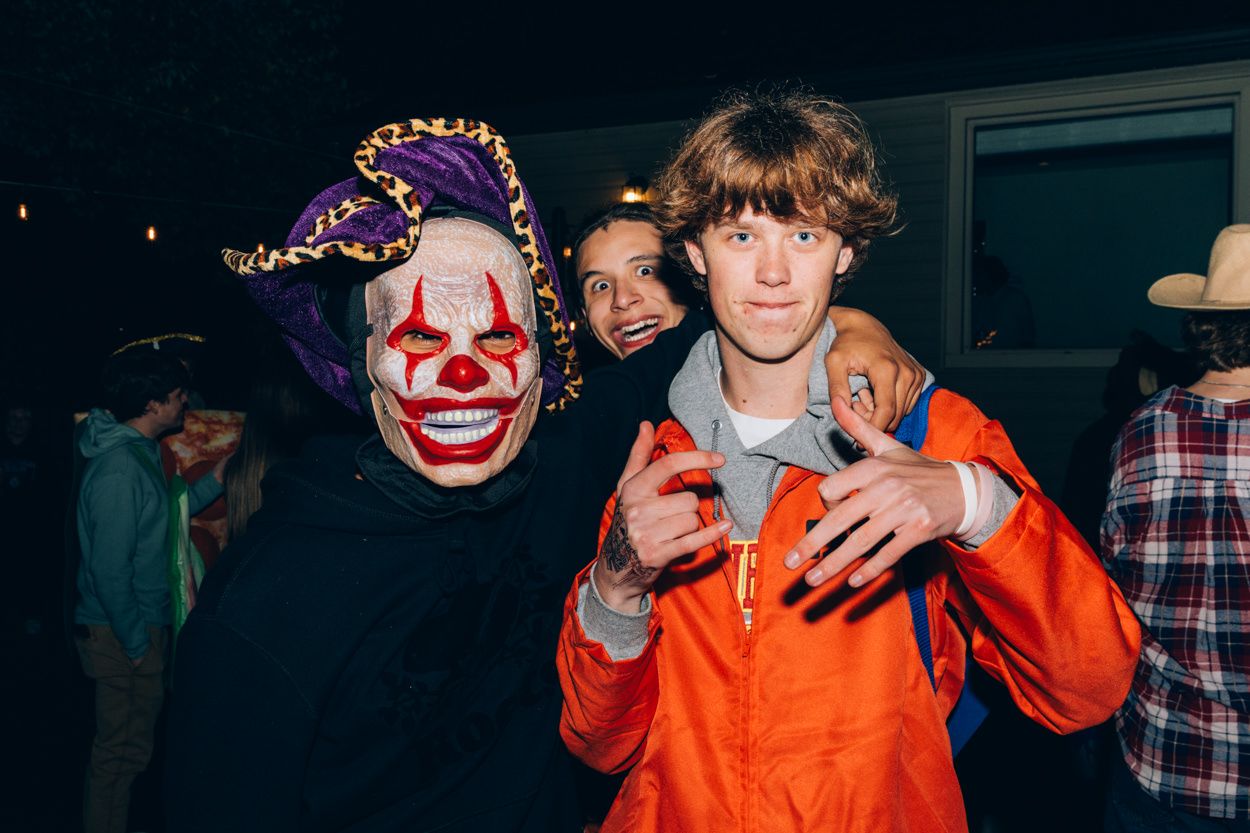 Person in clown mask and another in orange jumpsuit pose at Halloween party.