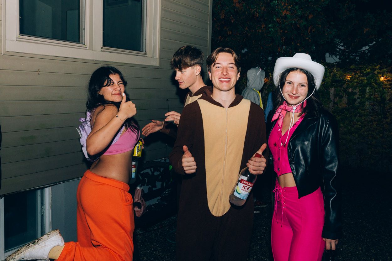 Friends in colorful costumes including a hot dog pose against a house exterior at night.
