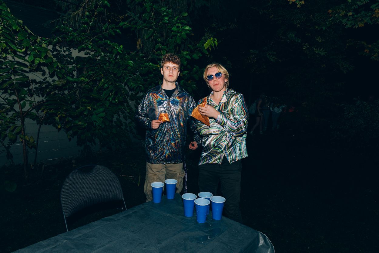 Two people in tie-dye outfits standing at a beer pong table with blue cups.