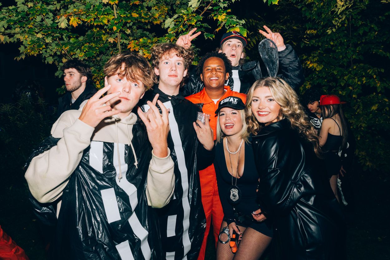 Party-goers in Halloween costumes pose together outdoors at night under tree branches.