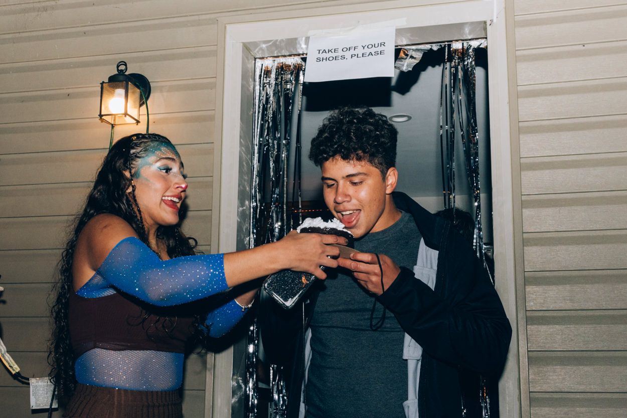 Two people playfully sharing a drink outside a house at night.
