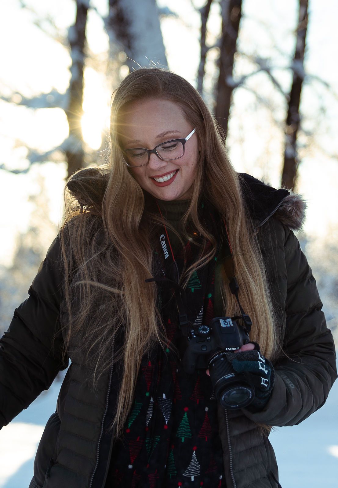 Winter portrait of someone wearing glasses and winter coat holding camera in snowy forest.