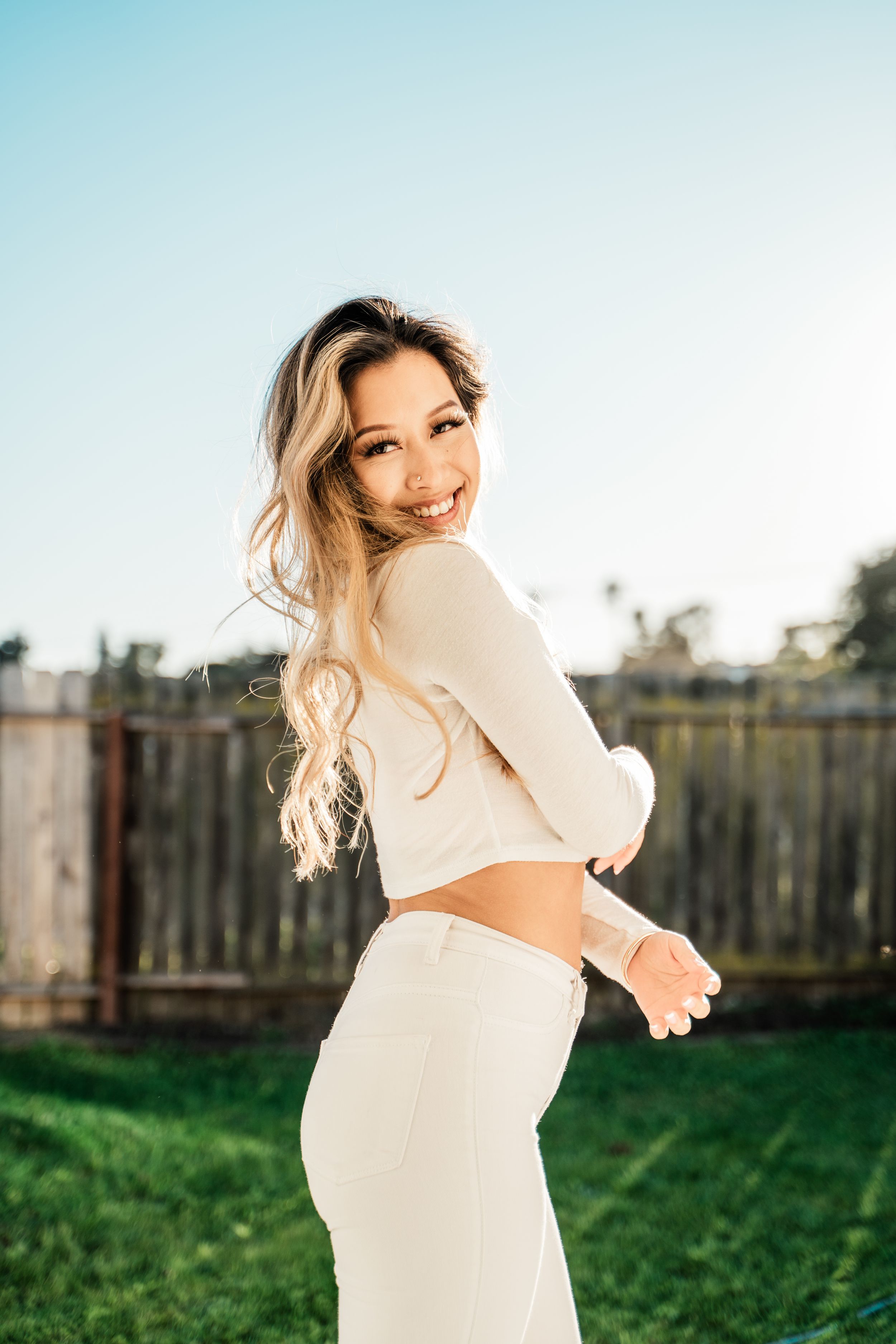 A smiling person wearing a white cropped sweater and matching pants poses in a backyard with green grass.