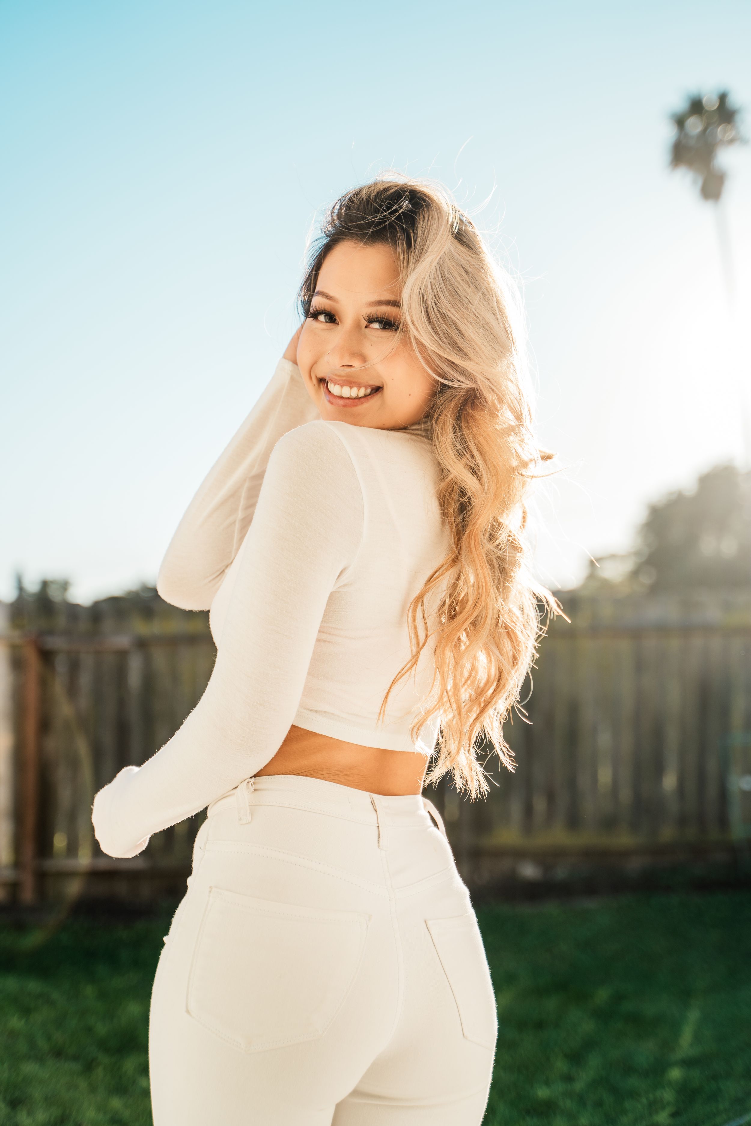 A person in a white crop top and matching pants poses outdoors on a sunny day with a palm tree in the background.