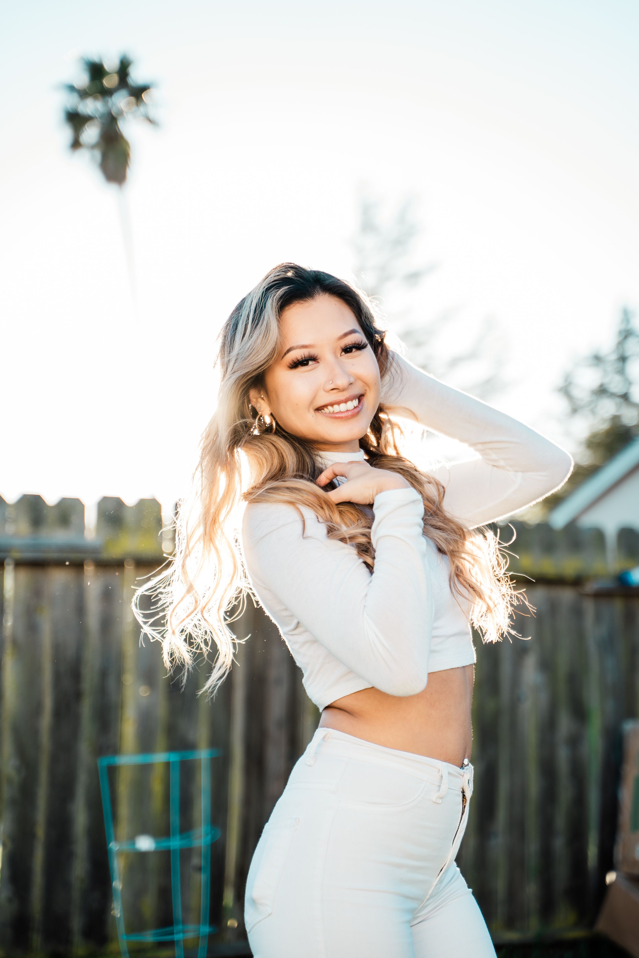 A young woman in white attire poses outdoors against a wooden fence with a palm tree in the background in soft lighting.