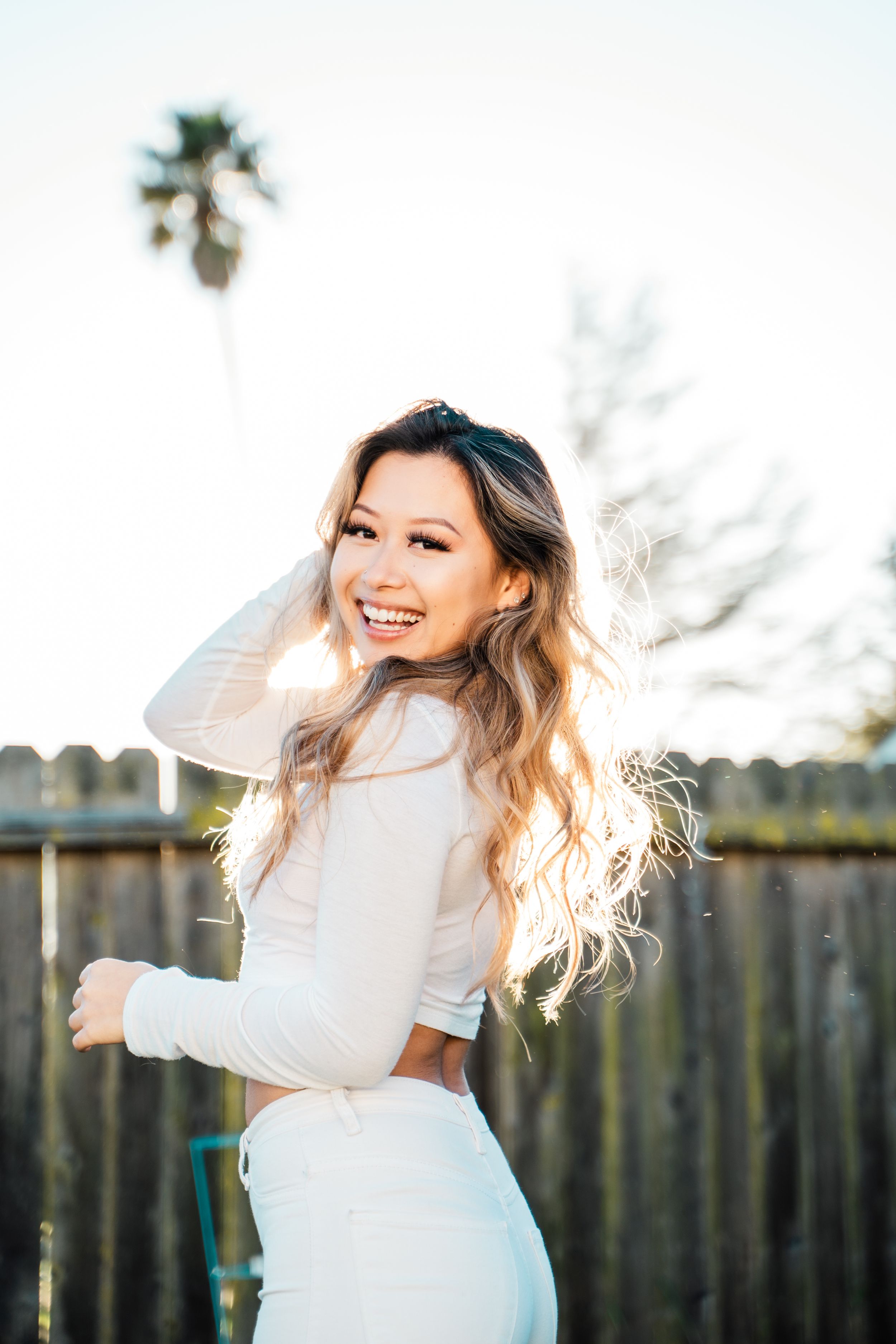 A woman in white outfit twirls joyfully in a backyard with afternoon sunlight.