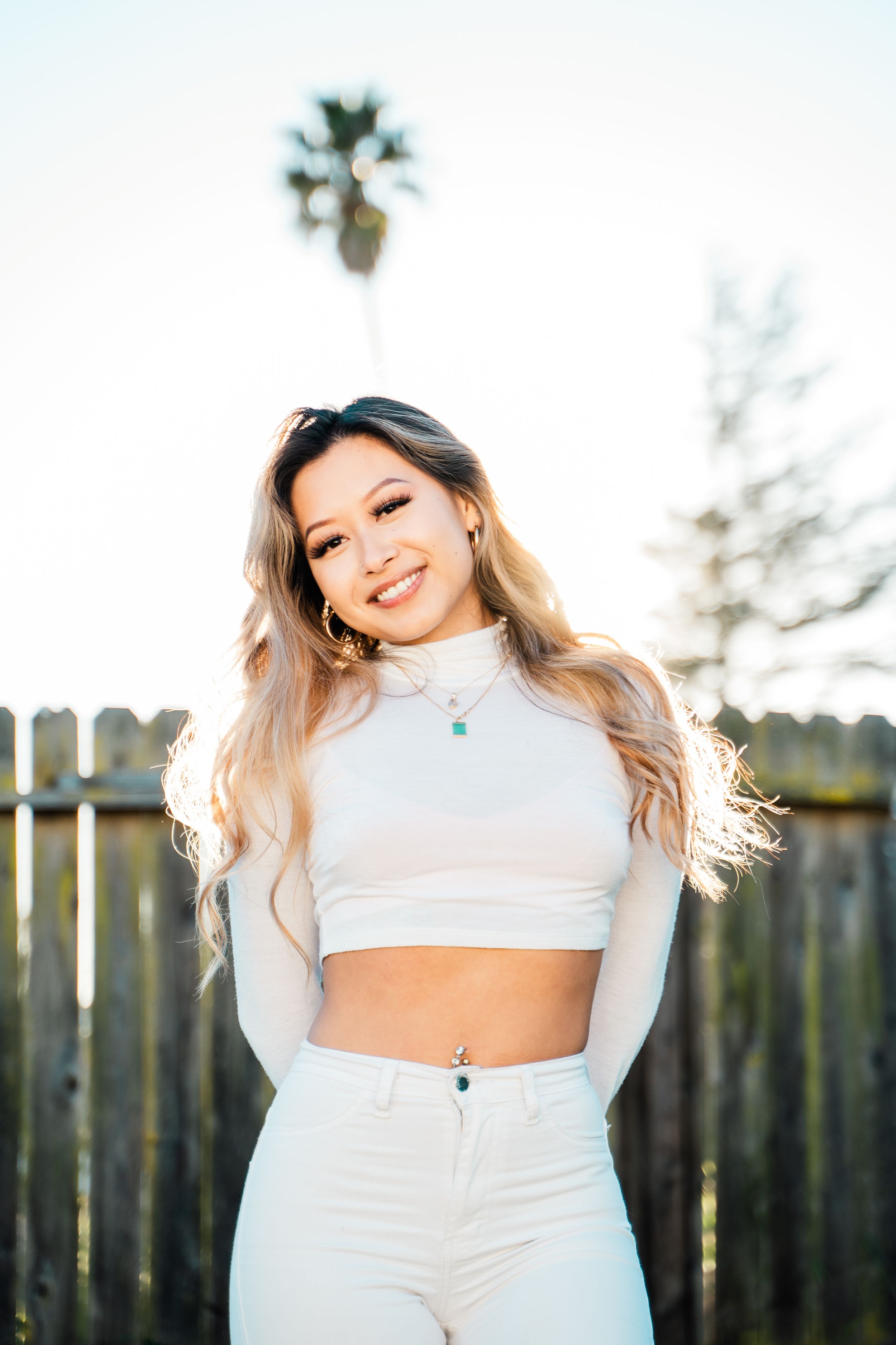 Sun-lit outdoor portrait of a person in a white crop top and high-waisted pants against a wooden fence.