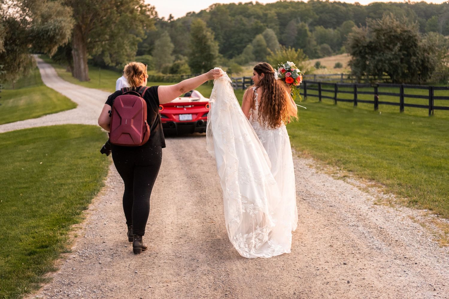 Two people walking down a gravel path with floral crowns and a vintage red car in the background.