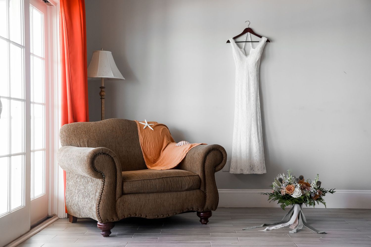 A wedding dress hangs on display next to a brown armchair and floral arrangement in a sunlit room.