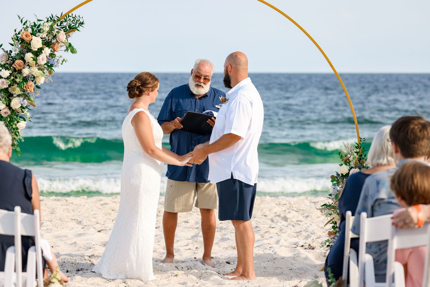 A beach wedding ceremony under a golden arch with waves in the background.