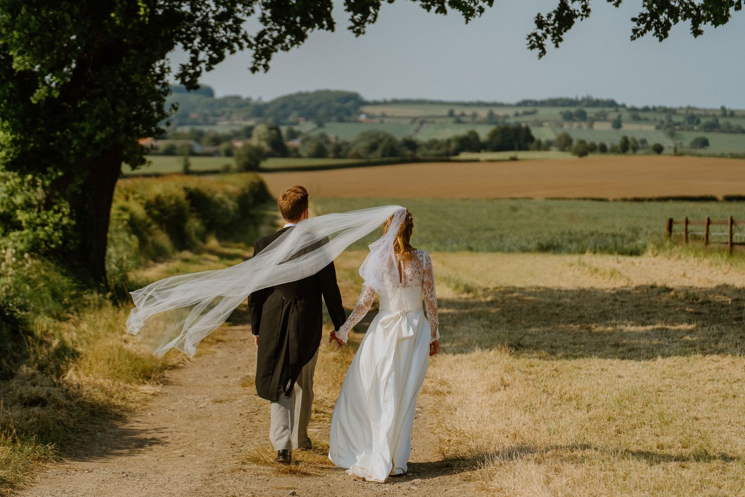 Couple walking along a countryside path with flowing white dress and veil catching the breeze.