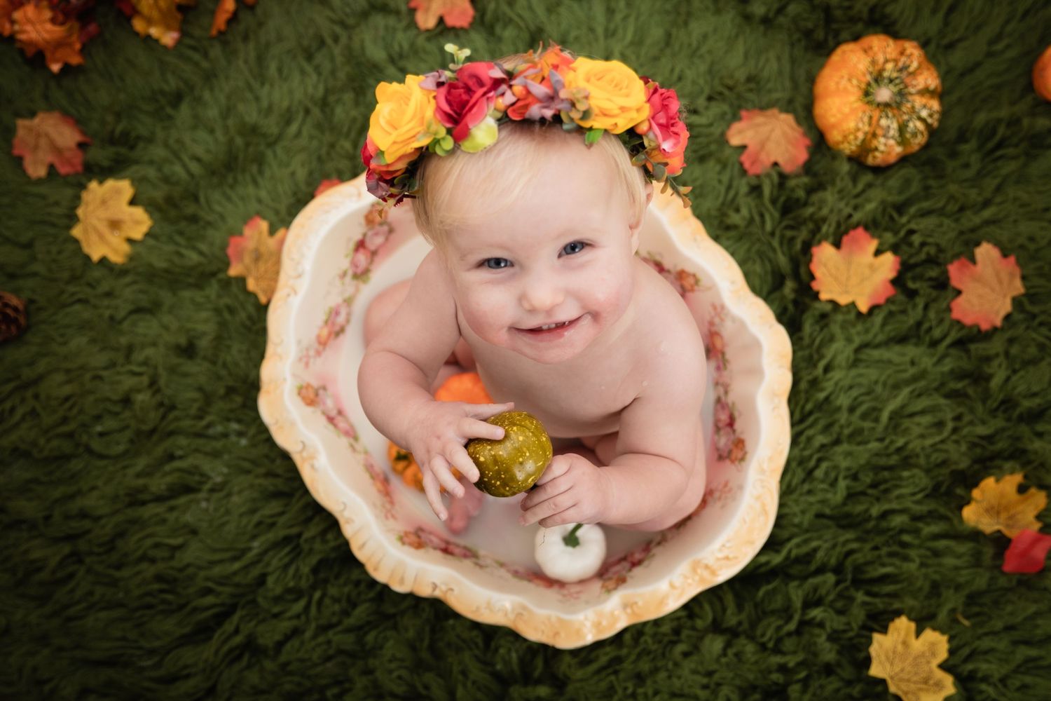 A smiling baby wearing a floral crown surrounded by autumn pumpkins and leaves.